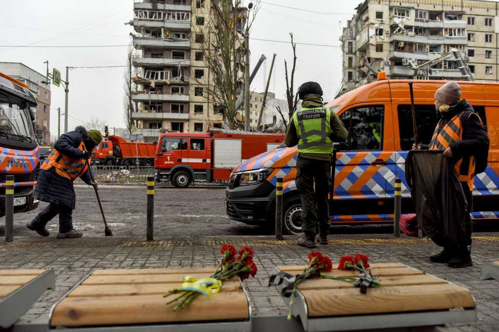 Dnipro (Ukraine), 15/01/2023.- Red flowers placed over the seats of a bus stop (front) as communal workers clean the street near the site of a damaged residential building (back) in Dnipro, southeastern Ukraine, 15 January 2023, amid Russia's invasion. At least 25 people died, including one child, 73 other were injured, including 13 children, and 43 reports of missing persons were received, after a rocket hit a nine-story building in Dnipro on 14 January, the State Emergency Service (SES) of Ukraine said in a statement. Russian troops entered Ukraine on 24 February 2022 starting a conflict that has provoked destruction and a humanitarian crisis. (Atentado, Rusia, Ucrania) EFE/EPA/OLEG PERTASYUK