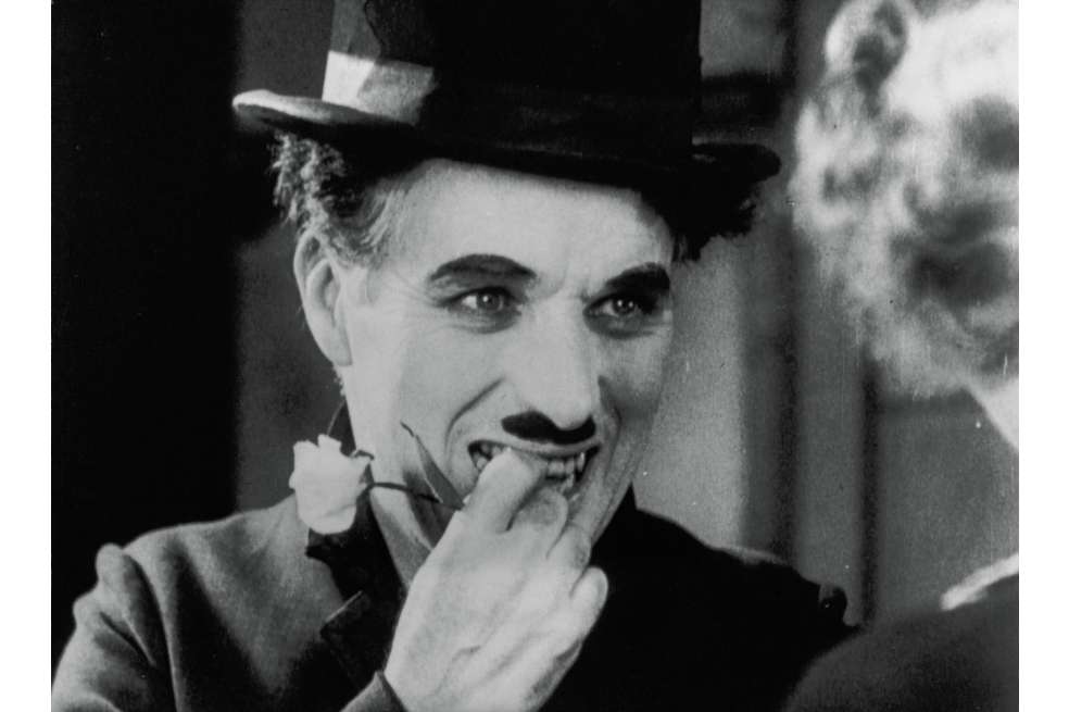 Primer plano de Charles Chaplin. La foto a blanco y negro muestra a Chaplin con un sombrero, su bigote insignia y los dedos de su mano derecha puestos sobre sus labios. Con ellos sostiene una flor.