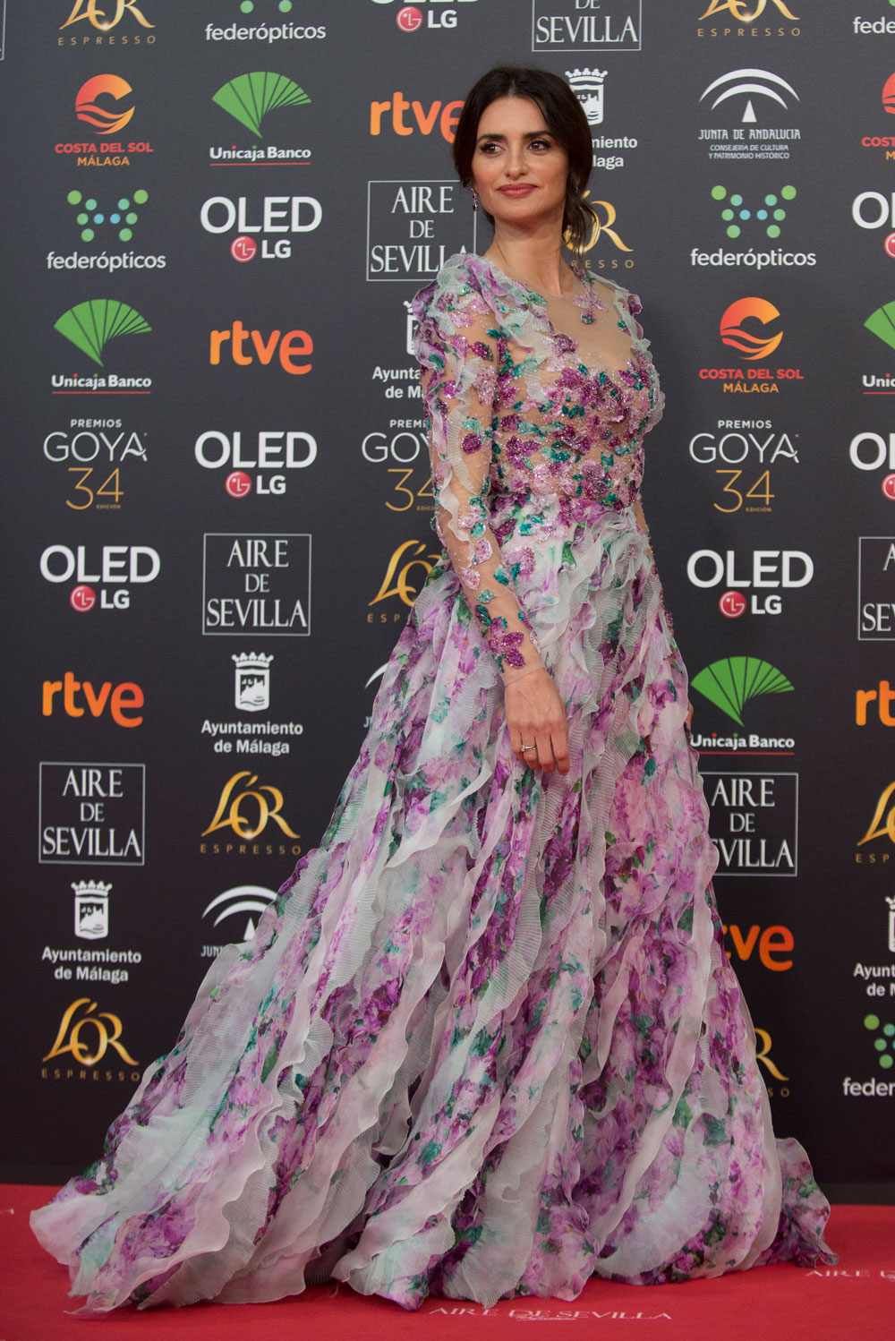 Este diseño multicolor, en tonos rosados, lilas y verdes ha causado auténtica sensación. El vestido de manga larga y tul semitransparente sobre el que los bordados de flores 3D cubren algunas partes del cuerpo, tiene una parte superior que se ciñe a la perfección sobre el torso. Desde la cintura caen en cascada tiras longitudinales de organza creando el efecto de volantes para así obtener una amplia falda de princesa que termina en cola.