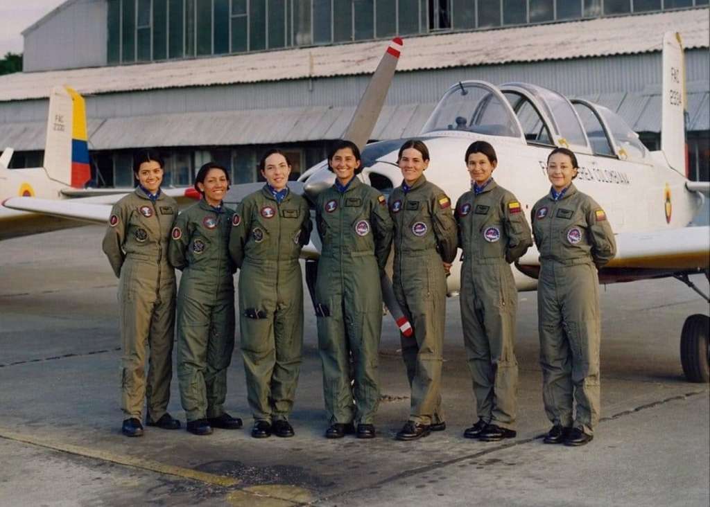 Grupo de siete mujeres con uniformes de vuelo verde oliva posan frente a una aeronave de la Fuerza Aérea Colombiana en un hangar. Todas llevan parches e insignias en sus trajes.