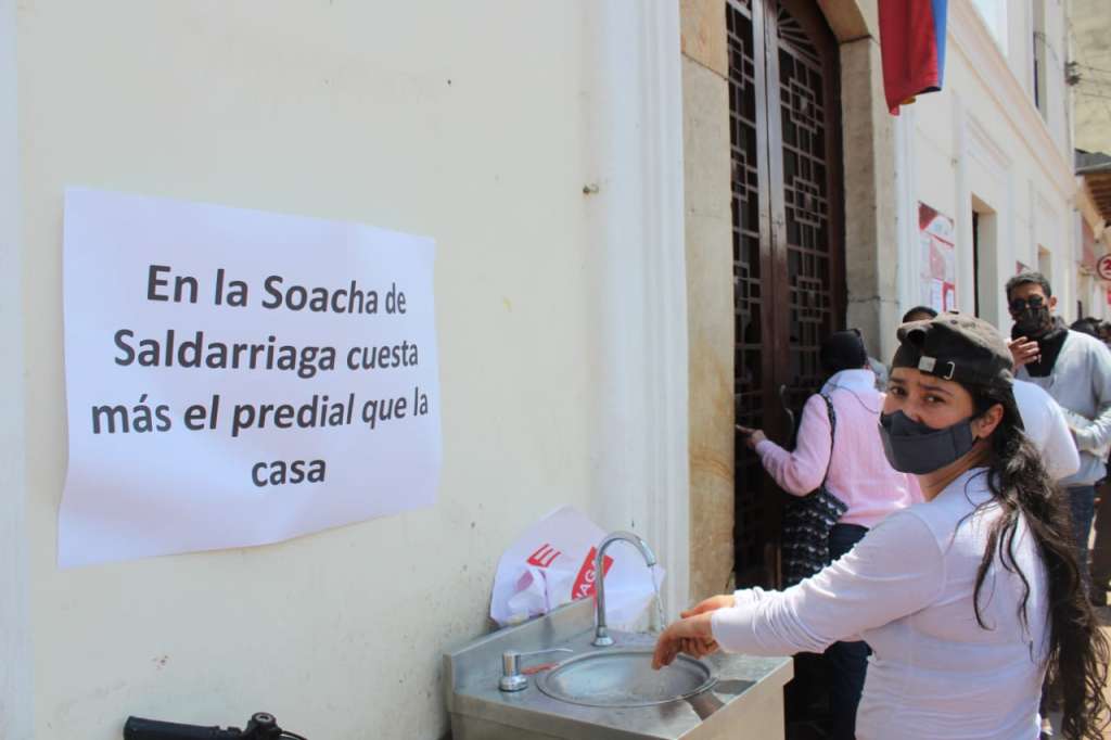 Una mujer se lava las manos junto a un cartel que dice: en la Soacha de Saldarriaga cuesta más el predial que la casa.