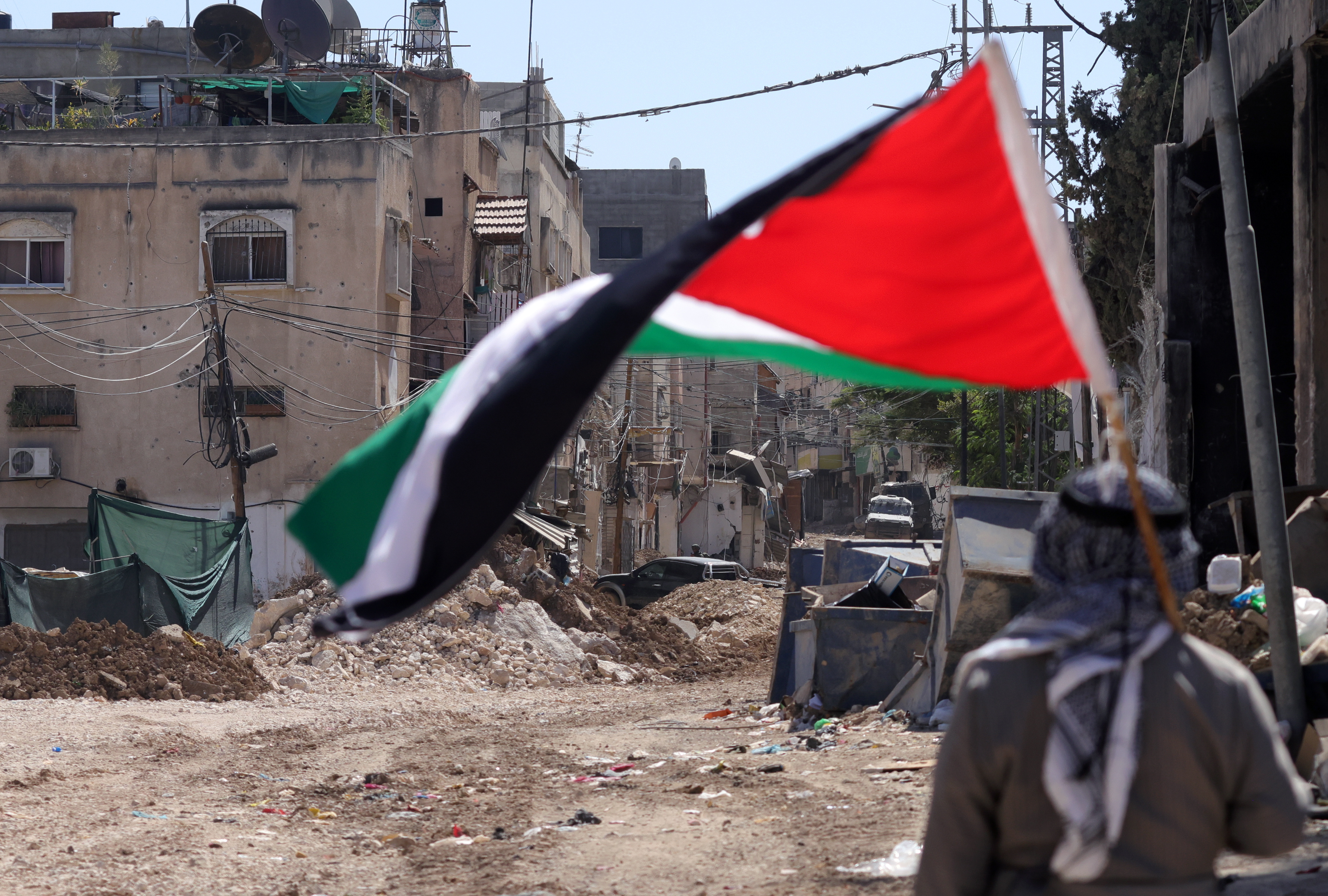 FOTODELDÍA Tulkarem (Cisjordania), 11/09/2024.- Un hombre sostiene una bandera palestina mientras soldados israelíes realizan una operación en el campo de refugiados de Tulkarem, cerca de la ciudad cisjordana de Tulkarem, este miércoles. Al menos dos palestinos murieron el 10 de septiembre durante las operaciones militares israelíes en Tulkarem y su campo de refugiados, según el Ministerio de Salud palestino. EFE/ Alaa Badarneh