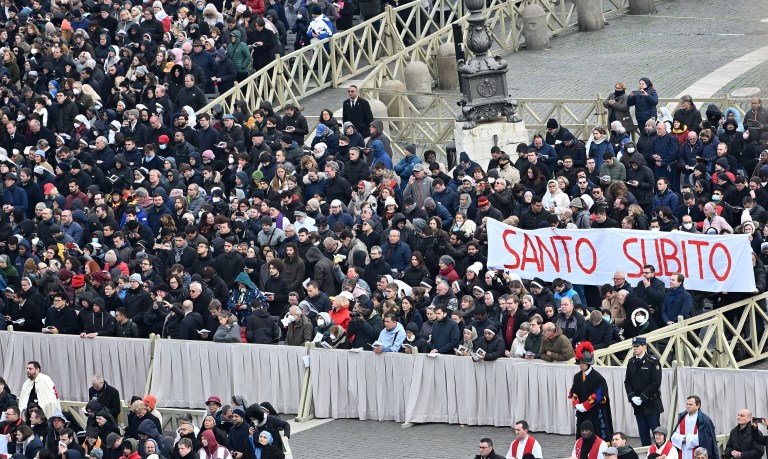 Los peregrinos sostienen una pancarta durante la ceremonia fúnebre del papa emérito que traduce, del italiano, "santo ahora".