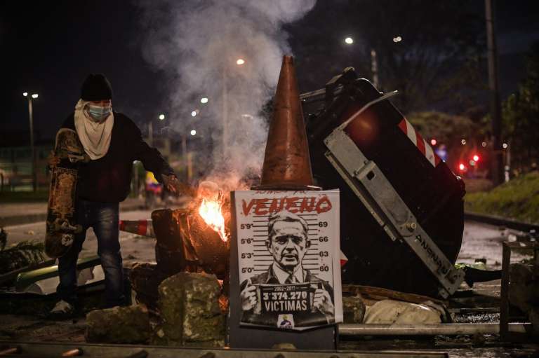 Un hombre al lado de un contenedor de basura incendiado donde hay un afiche contra Álvaro Uribe.
