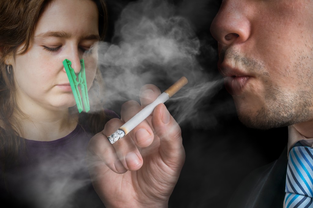 Passive smoking concept. Selfish man is smoking a cigarette. Woman cant breathe and has peg on her nose.