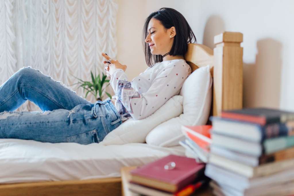 Young women in casual clothing, lying down in bad and using phone. It's time to gether the people, weekend is just started.