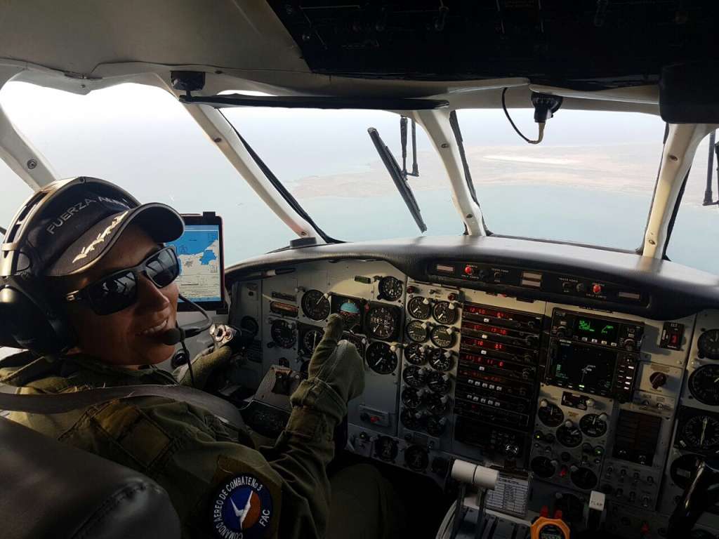 Cabina de avión en vuelo. Una piloto con gafas oscuras, gorra de la Fuerza Aérea y uniforme con el logo del Comando Aéreo de Combate No. 3 opera los controles. Se observan instrumentos analógicos y una tableta con mapa de ruta; al fondo, paisaje costero visto desde el aire.