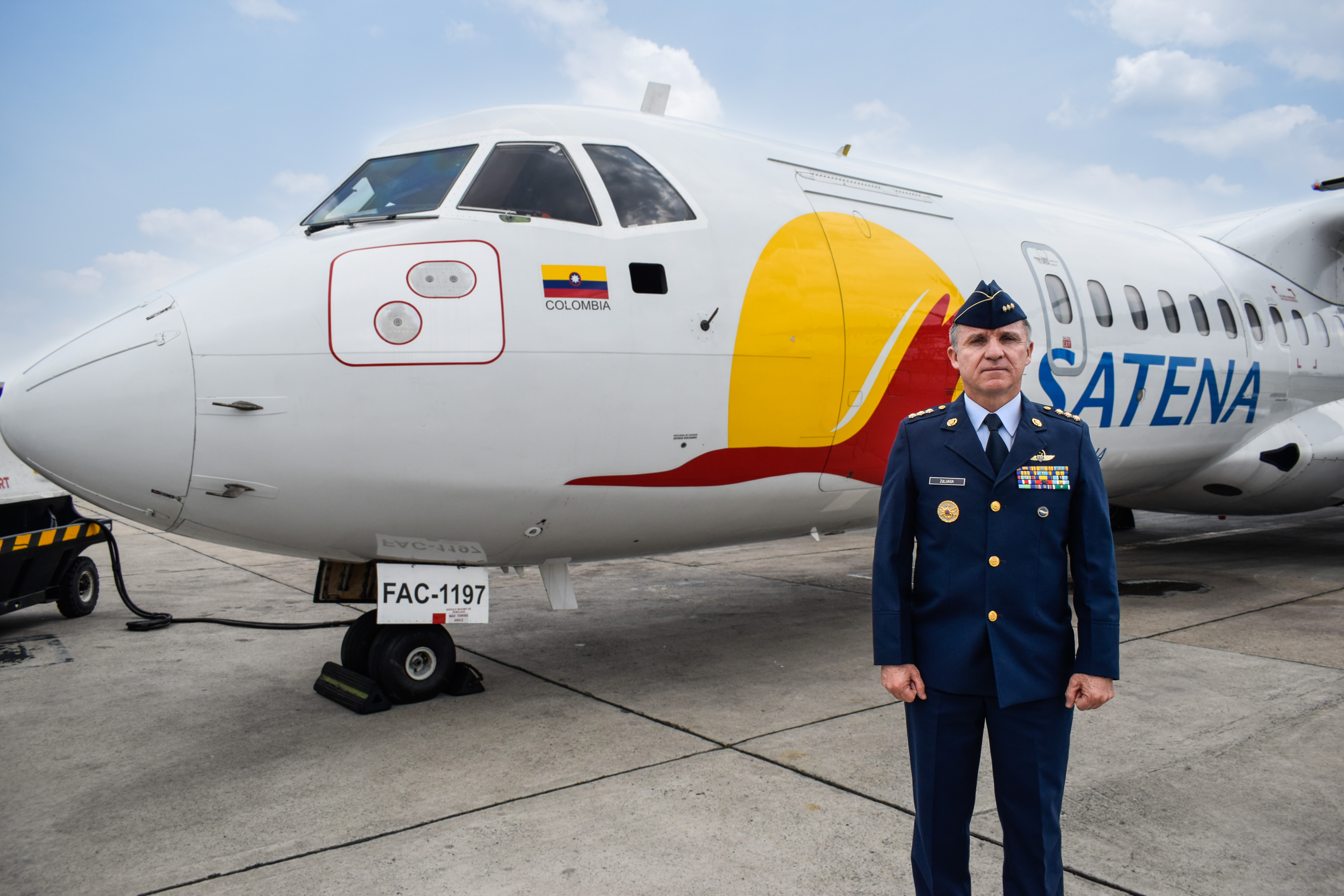 Oficial de la Fuerza Aérea junto a avión Satena en pista, con bandera de Colombia visible en el fuselaje.