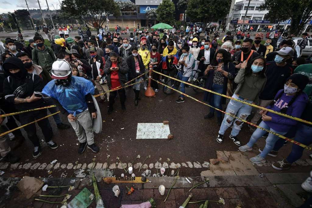 Varias personas rodean una zona cercada con cintas de no pase en las que hay velas y flores