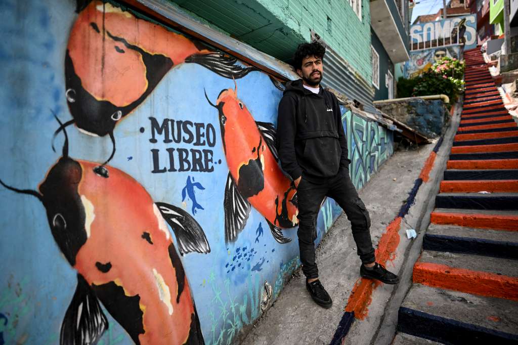 Colombian artist Wilson Ni�os, member of the SurVamos collective, poses for a picture in front of a mural during the free museum event at the Ciudad Bolivar neighbourhood in Bogota, on December 22, 2022. - A gigantic eye guards the Bogota neighborhood of Ciudad Bolivar. Realistic and painted in grayscale, the mural stands as a challenge against the government initiative that painted the facades of the most stigmatized area of the Colombian capital in bright colors. "It is a discriminatory program, because it was not carried out in all parts of the city, it was only for some poor neighborhoods", Wilson Ni�o of the SURVAMOS collective, who paints his own murals in Ciudad Bol�var as an act of "resistance" against local government interventions, told AFP. (Photo by Juan BARRETO / AFP)