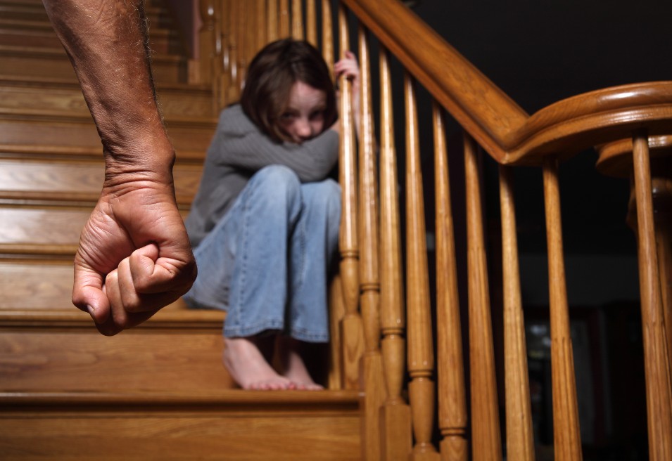 Una niña atemorizada se aferra a las barandas de una escalera de madera mientras se padre la amaneza con el puño cerrado.