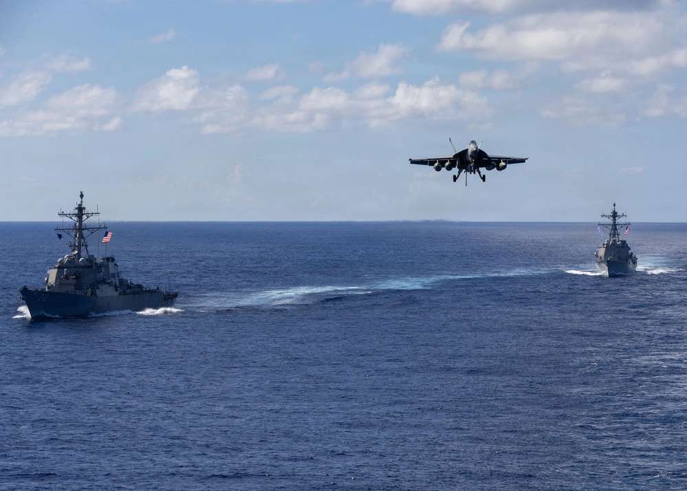 Dos destructores de misiles guiados navegan en formación mientras un F/A-18 Super Hornet, del USS Gerald R. Ford, desciende para aterrizar en el portaaviones.