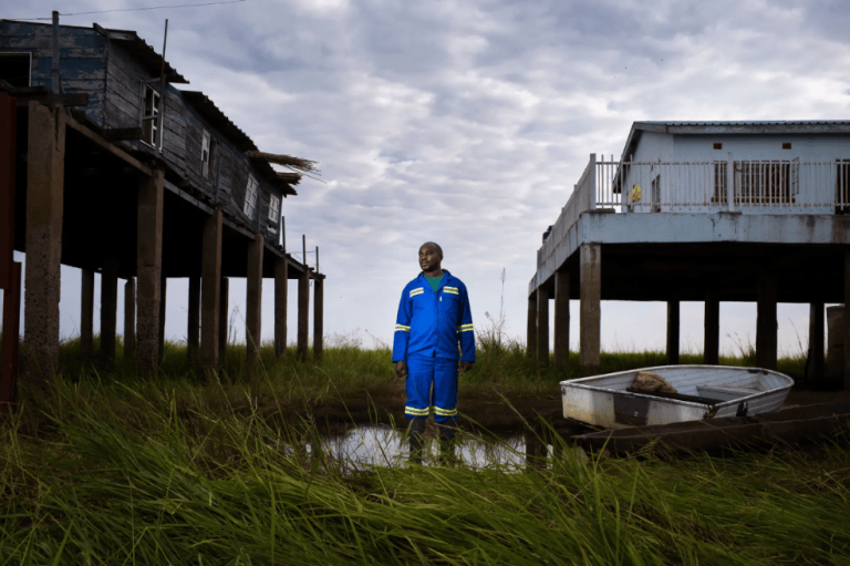 Las inundaciones perdidas: Un retrato de Lubinda Lubinda, jefe de estación de la Autoridad del Río Zambeze en Zambia. El bajo nivel de las aguas hace que su nueva casa (a la derecha) no tenga que ser construida tan alta. El pueblo barotse de la llanura aluvial está sometido a sequías más frecuentes debido al cambio climático y la deforestación.