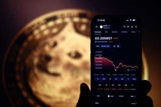 A photo illustration created in London on February 6, 2025, shows a smartphone displaying information on the price fluctuations of Dogecoin, backdropped by the doge meme displayed on the screen of a computer screen.