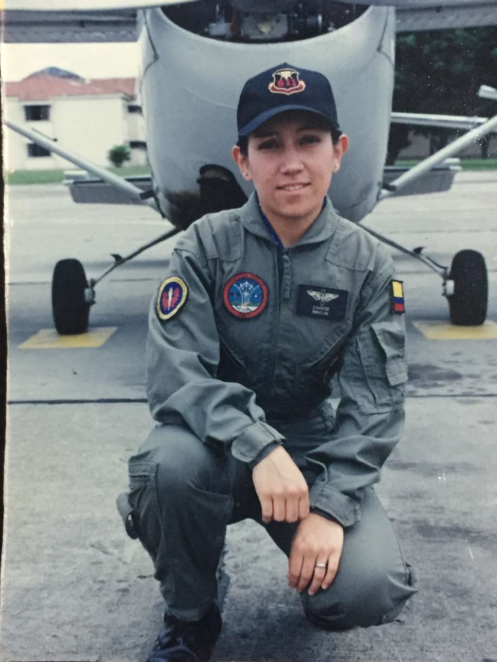 Cadete con uniforme de vuelo verde oliva arrodillada en la pista frente a un avión de instrucción con hélice. Lleva gorra azul con insignia y parches en el pecho, entre ellos la placa con su nombre.