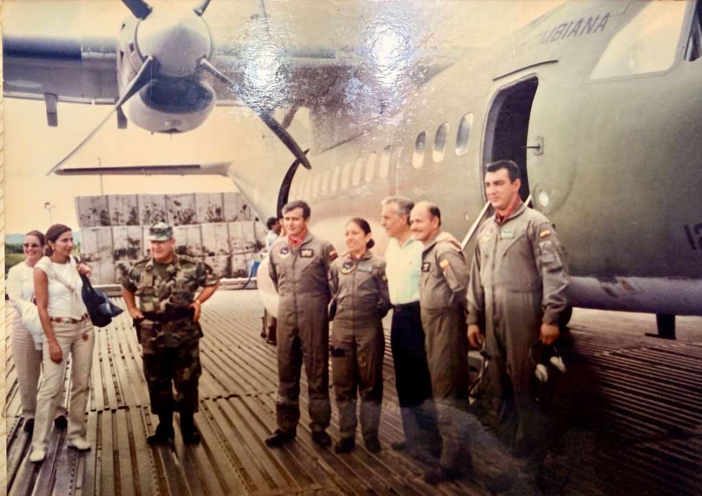 Grupo de personas sobre una plataforma metálica junto a un avión militar gris de la Fuerza Aérea Colombiana. Entre ellos, pilotos con uniformes de vuelo, un militar en camuflado y civiles. Magda, la mujer más pequeña, está ubicada aproximadamente en el centro del grupo.