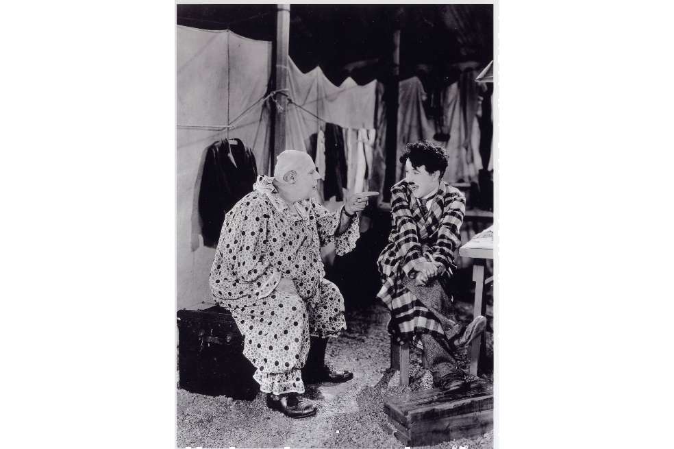Charles Chaplin está sentado hablando con un personaje del circo. Chaplin tiene puesto un traje de cuadros.