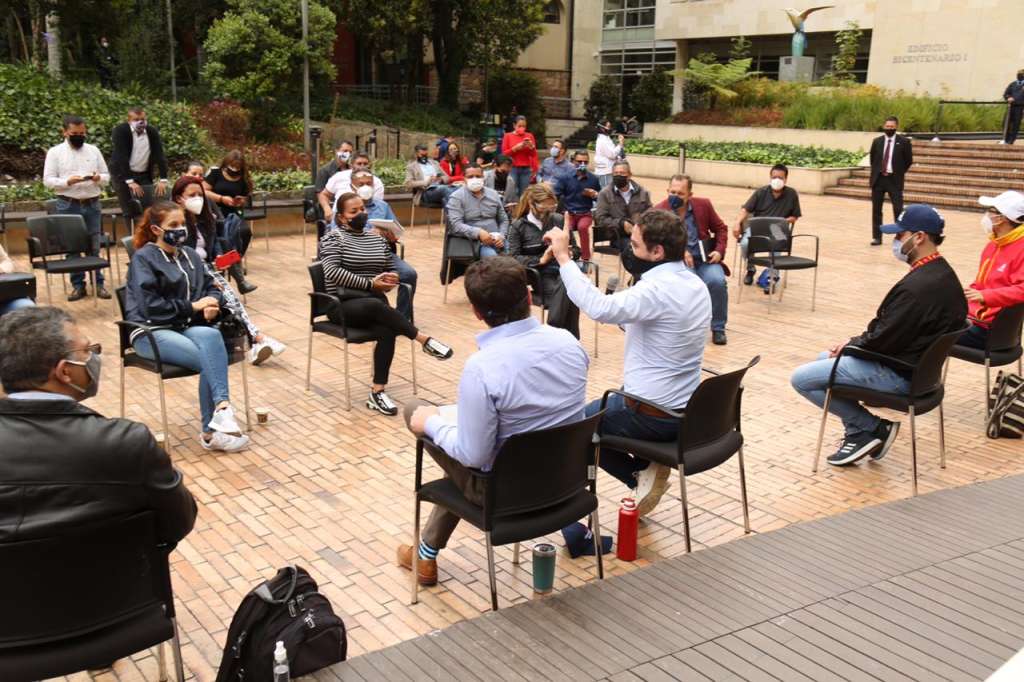 Varias personas se encuentran reunidas y sentadas en una plazoleta.