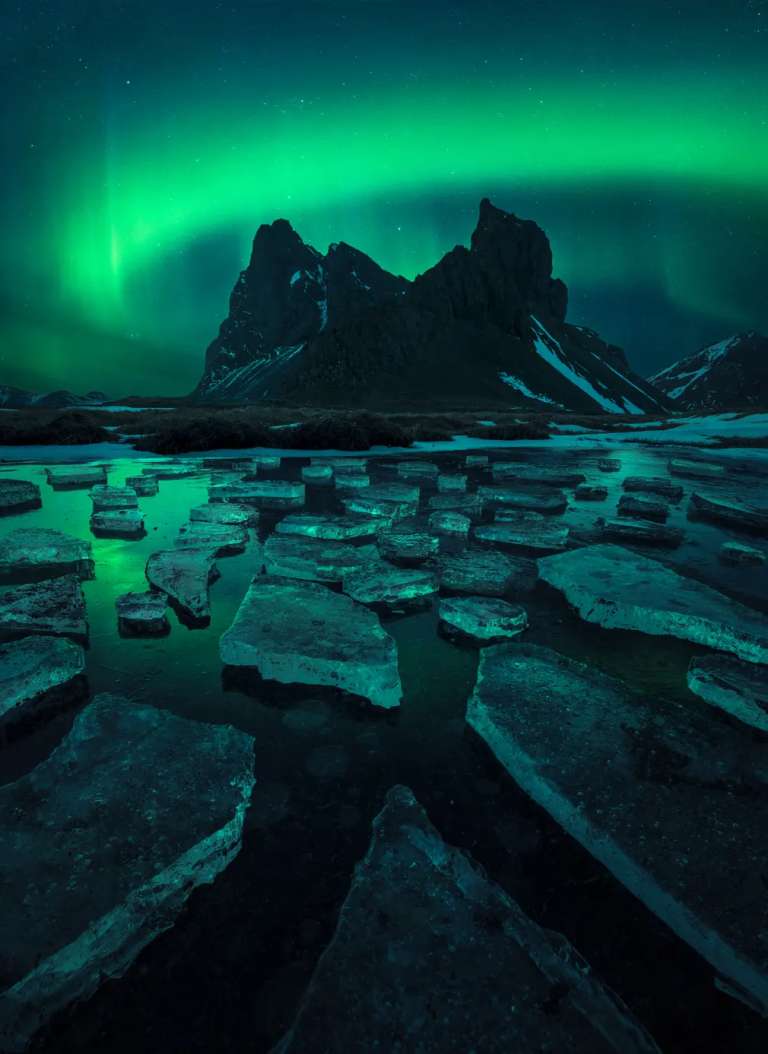 La imagen muestra la aurora boreal bailando, reflejada en un pequeño lago helado sobre la montaña Eystrahorn.