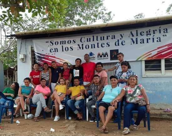 Foto de un grupo de hombres y mujeres de la población LGBT, sentados y de pie. Están en un entorno rural y detrás de ellos hay una pancarta que dice: "Somos cultura y alegría en los Montes de María".