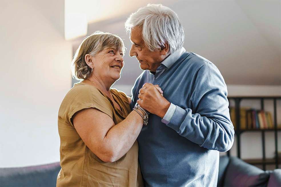 Grandmother and grandfather carefree dancing at home holding hands and looking on each other eye - Elderly couple celebrates their anniversary at home