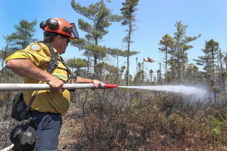 Esta imagen del 1 de junio de 2023, cortesía del Gobierno de Nueva Escocia en Canadá, muestra al bombero Walter Scott rociando el suelo mientras un bombardero de agua vuela para echar una carga de agua sobre el fuego, en Barrington Lake, Canadá.