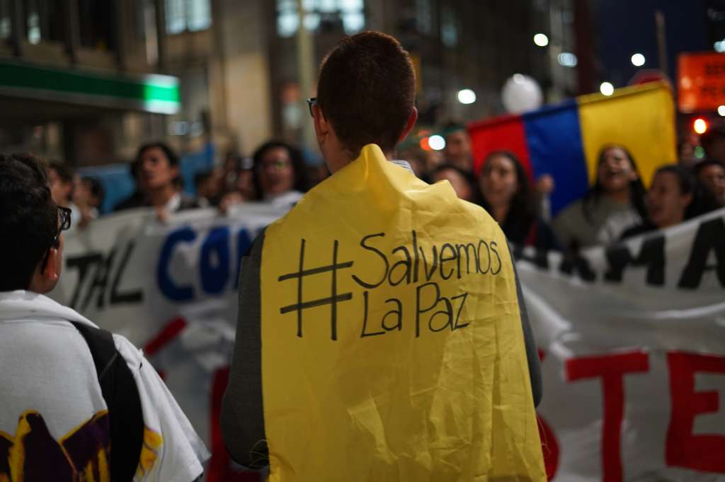 En la foto hay un joven de espaldas usando una bandera como una capa de superhéroe en la que dice "Salvemos la Paz". En el fondo se ven a jóvenes en una marcha. Días después del resultado del plebiscito por la paz, miles de ciudadanos salieron a marchar desde diferentes lugares del país exigiendo que se acordara lo necesario para continuar con el Acuerdo. / Foto: Diego Cuevas.