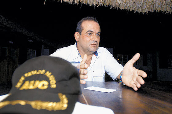 Salvatore Mancuso está sentado, con las manos abiertas y al lado hay una gorra con la palabra AUC.