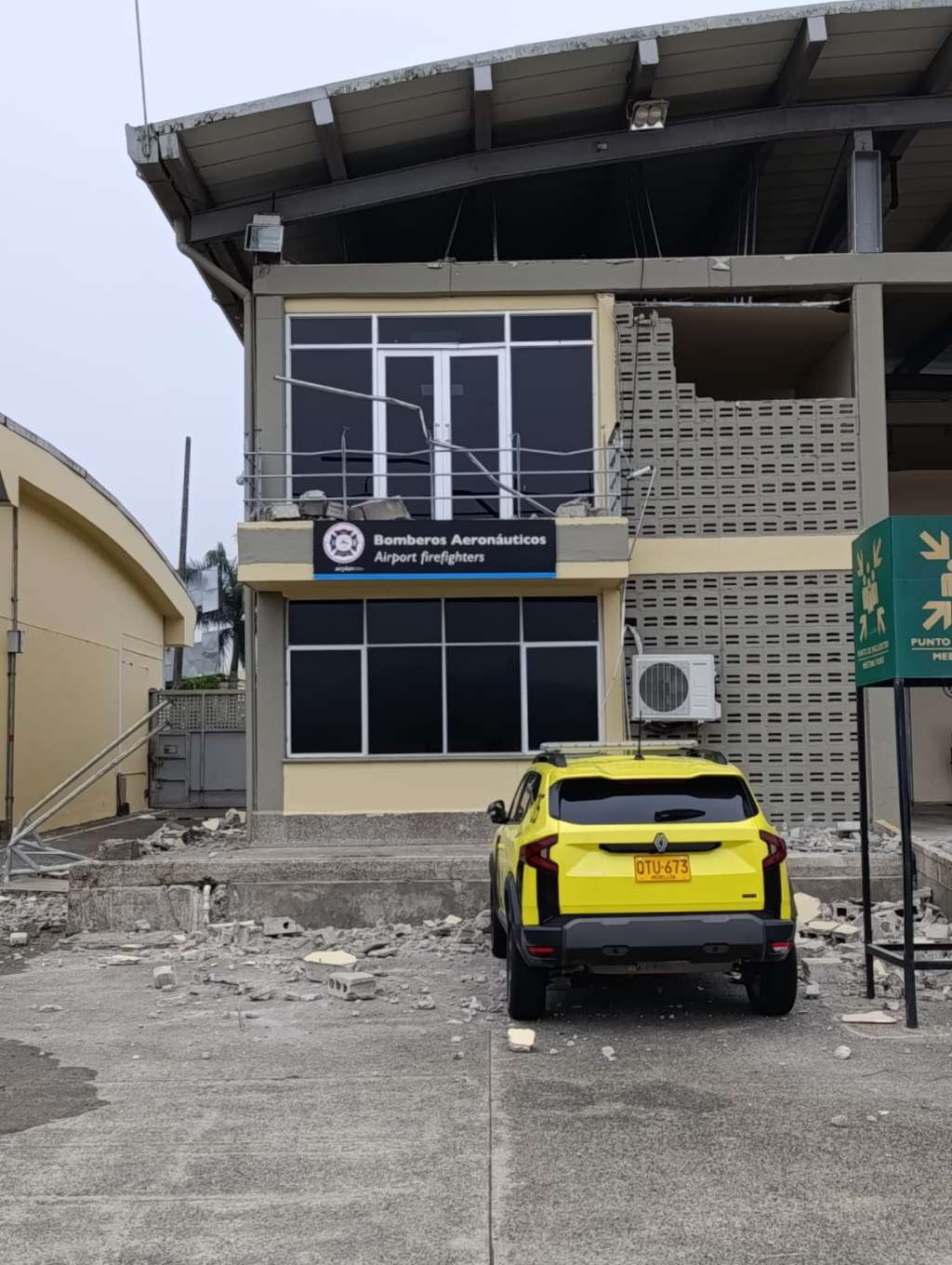 Edificio de dos pisos de los Bomberos Aeronáuticos con daños estructurales visibles: ventanas rotas en la parte superior, baranda metálica doblada y grietas en las paredes. En el suelo hay escombros y fragmentos de concreto frente a la entrada. Un automóvil amarillo con placa OTU-673 está estacionado junto a la fachada, rodeado de restos caídos. La escena refleja un inmueble de servicios de emergencia afectado por colapso parcial o evento sísmico.
