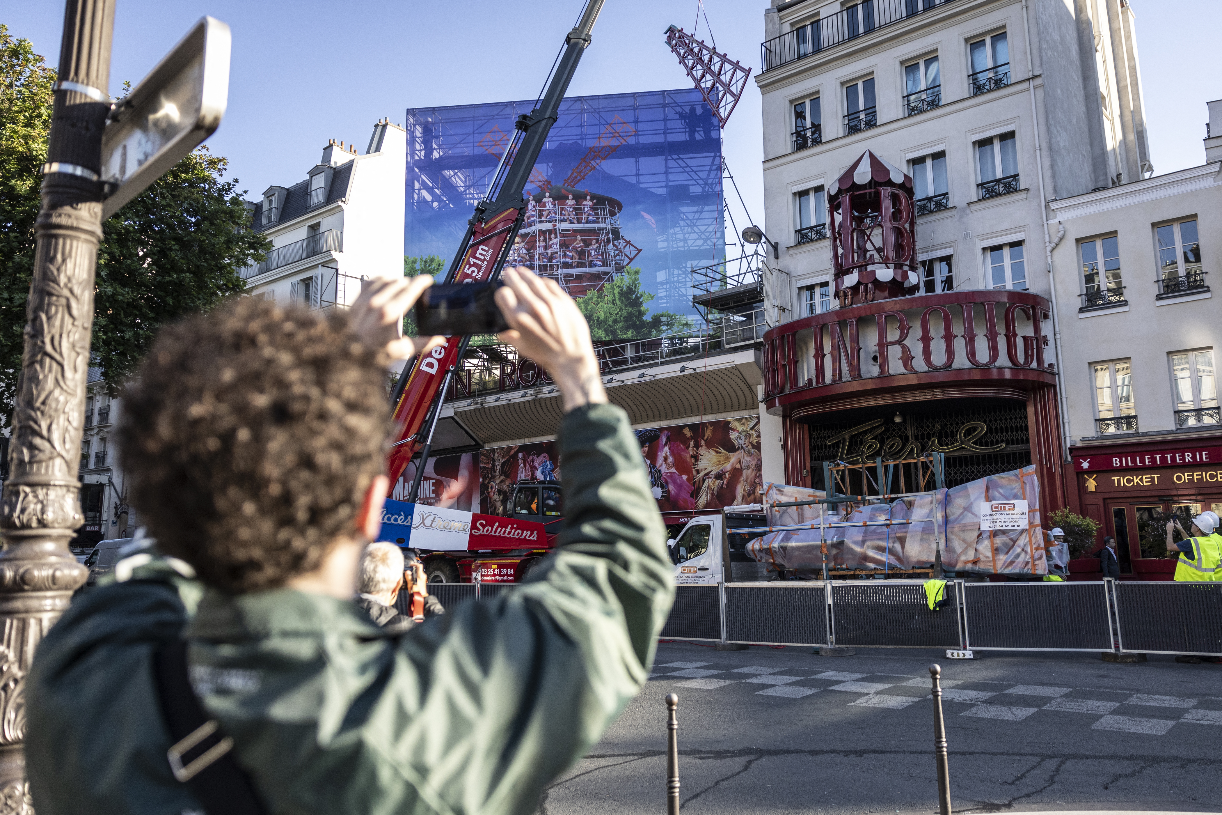 A pedestrian takes a picture of a crane lifting one of the four new windmill sails to install it on the top of the Moulin Rouge cabaret following the collapse of the former ones, in Paris on June 24, 2024.