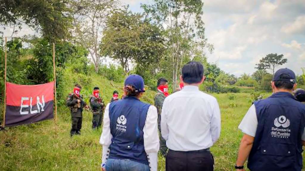 Las personas en la fotografía se encuentran en un entorno natural, con arboles y pasto.
En la parte izquierda de la imagen, se encuentran soldados de la guerrilla ELN, con su uniforme camuflado y sus distinciones rojas con negro. Además, mantienen en pie una bandera de los mismos colores y con las letras "ELN" en blanco.
Por otro lado, se encuentran tres personas de espaldas, dos de ellas tienen chalecos de la defensoría del pueblo y el tercer sujeto, estaría de camisa blanca, presuntamente como un representante de la Iglesia Católica