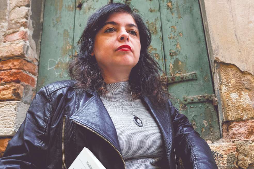 Mariana Enríquez, escritora argentina, tiene chaqueta de cuero color negro, blusa gris y un pendiente que cae sobre su pecho. Tiene el pelo suelto y lo labios pintados de rojo.